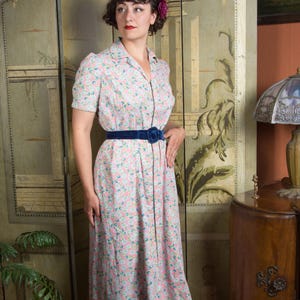 May include: A midi-length dress featuring a light pink background with a dense pattern of small blue and white flowers. The dress has short sleeves, a collared neckline, and a dark blue velvet belt with a circular buckle at the waist.