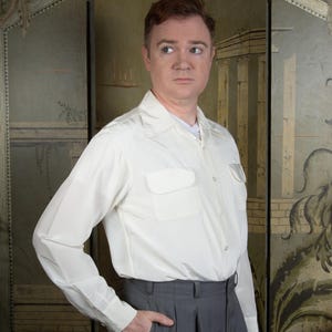 May include: A cream-colored button-down shirt with two chest pockets and gray pleated trousers. The shirt has long sleeves and a classic collar. The trousers have a high waist and a relaxed fit.