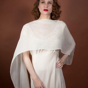 May include: A cream-colored vintage dress with a matching sheer shawl. The dress has a simple, elegant design, and the shawl drapes over the shoulders. The model has red lipstick and curly brown hair. The background is a solid brown.