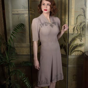 Peut inclure: Robe taupe à manches courtes, longueur genou, ornée de broderies au col et à l'ourlet. La modèle porte des talons gris. Le fond comprend un paravent décoratif et une plante en pot. Style vintage classique.
