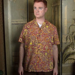 Peut inclure: Une chemise à manches courtes avec un motif cachemire audacieux en rouge, jaune et blanc. La chemise est portée sur un t-shirt blanc et associée à un pantalon marron. Le style suggère une esthétique rétro ou vintage.