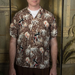 Könnte beinhalten: Braunes kurzärmeliges Hemd mit weißem Unterhemd. Das Hemd hat ein sich wiederholendes Muster aus Figuren, Palmen und Fackeln. Das Hemd hat einen klassischen Camp-Kragen.