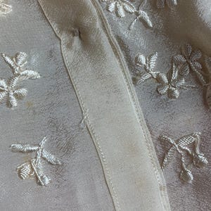 Puede incluir: Primer plano de una tela color crema con patrones florales bordados. La tela tiene un brillo satinado y una textura delicada. El bordado es en un color crema a juego, creando un dise&ntilde;o sutil y elegante.