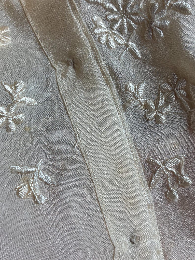 Puede incluir: Primer plano de una tela color crema con patrones florales bordados. La tela tiene un brillo satinado y una textura delicada. El bordado es en un color crema a juego, creando un dise&ntilde;o sutil y elegante.