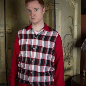 Può includere: Una camicia a quadri rossi e neri con maniche rosse. La camicia ha un motivo a quadri bianchi, neri e rossi sul davanti e un colletto rosso. La persona nell'immagine indossa una maglietta bianca e pantaloni grigi.