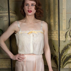 Puede incluir: Camisola color crema con detalles bordados y lazo amarillo, combinada con pantalones de cintura alta color melocot&oacute;n. El conjunto recuerda a lencer&iacute;a o ropa de dormir vintage. La modelo est&aacute; de pie frente a una pantalla decorativa.