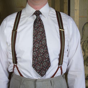May include: A white dress shirt with a floral patterned tie and brown suspenders. The suspenders have a brown and green stripe design and gold-colored clasps. The tie features a repeating pattern of red and gray roses.