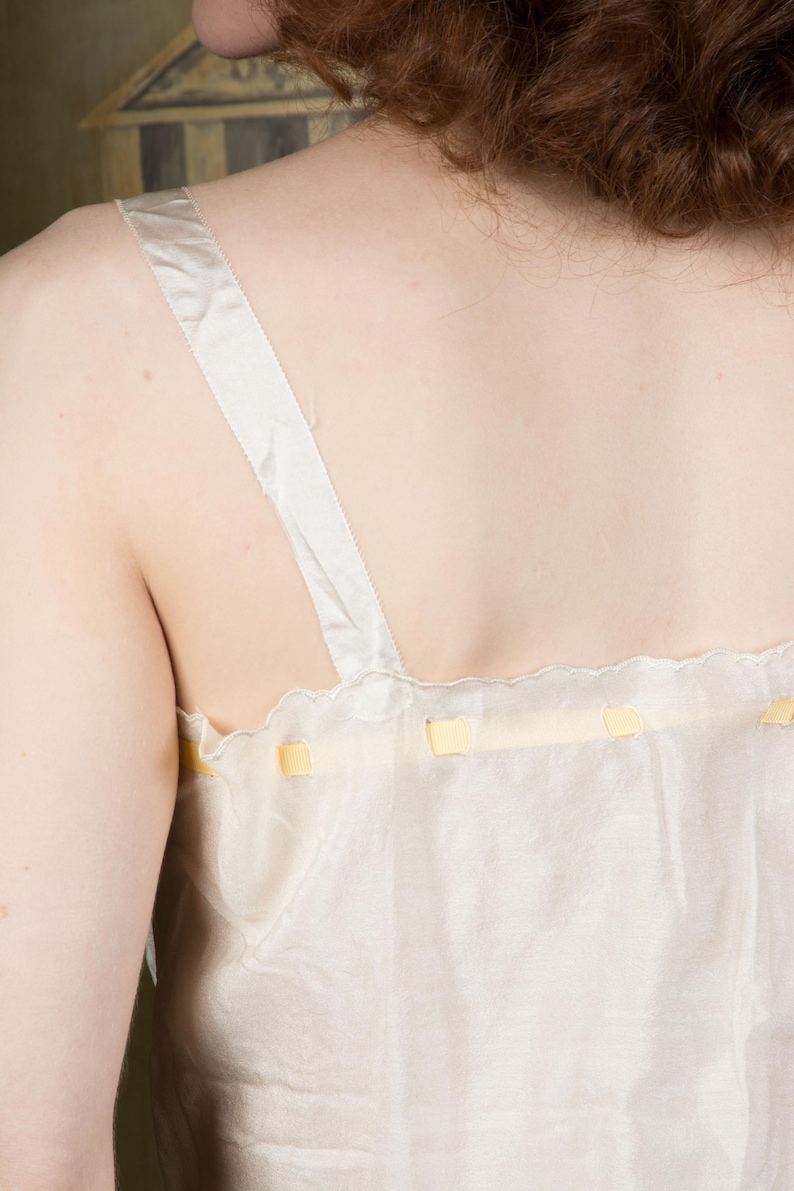Puede incluir: Camisola color crema con delicados lazos amarillos a lo largo del escote. La camisola presenta finas correas de sat&eacute;n blanco. La tela parece transparente y ligera, lo que sugiere un estilo vintage o de lencer&iacute;a.
