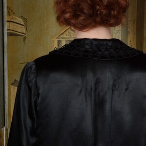Op de afbeelding: Een zwarte satijnen robe met een decoratieve kraag. De kraag heeft een ingewikkeld, donker geborduurd ontwerp. De robe heeft lange mouwen en een gladde, glanzende textuur. De achtergrond toont een glimp van een muur in vintage stijl.
