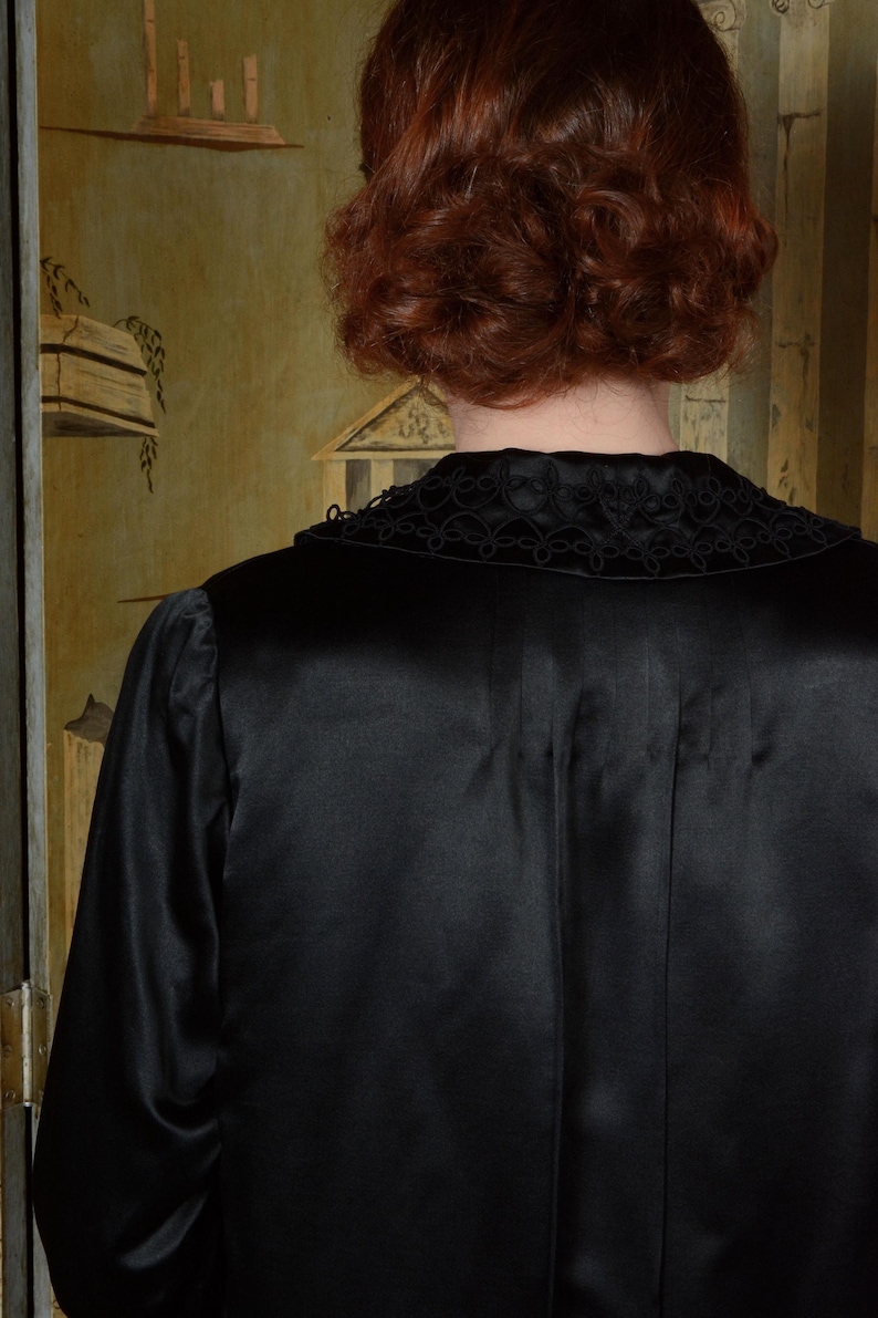 Op de afbeelding: Een zwarte satijnen robe met een decoratieve kraag. De kraag heeft een ingewikkeld, donker geborduurd ontwerp. De robe heeft lange mouwen en een gladde, glanzende textuur. De achtergrond toont een glimp van een muur in vintage stijl.