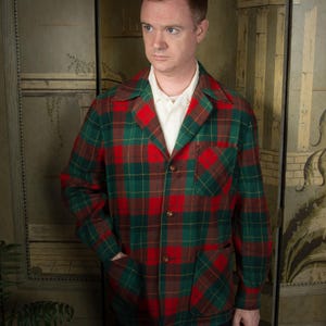 Puede incluir: Chaqueta a cuadros verdes, rojos y marrones con cuello con solapas, bolsillos de parche y cierre de botones. La chaqueta se lleva sobre una camisa blanca con cuello. La persona lleva pantalones negros.