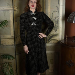 Puede incluir: Un vestido negro de estilo vintage con un sutil estampado blanco. El vestido presenta mangas largas, cuello alto con lazos decorativos y falda midi. El vestido se lleva con zapatos negros.
