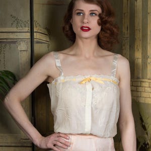 Puede incluir: Conjunto de lencer&iacute;a color crema con una parte superior transparente con delicados detalles de encaje y un lazo amarillo. La parte superior tiene tirantes de sat&eacute;n. La modelo lleva una parte inferior rosa claro a juego. El fondo incluye un biombo decorativo.