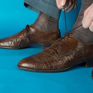 May include: Brown leather dress shoes with a textured, crocodile-like pattern. The shoes have laces and a low heel. The image shows a person tying the laces of one shoe. The background is a solid blue.