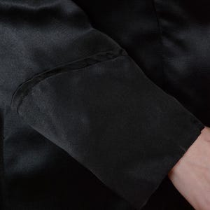 Op de afbeelding: Close-up van een zwarte satijnen mouw. De mouw heeft een op maat gemaakte manchet met subtiele naaddetails. De stof lijkt glad en heeft een lichte glans, wat wijst op een formeel kledingstuk.