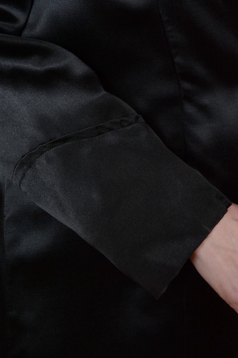 Op de afbeelding: Close-up van een zwarte satijnen mouw. De mouw heeft een op maat gemaakte manchet met subtiele naaddetails. De stof lijkt glad en heeft een lichte glans, wat wijst op een formeel kledingstuk.