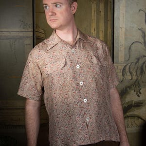 May include: A short-sleeved button-down shirt with a beige and red paisley pattern. The shirt has a camp collar and a single chest pocket. The buttons are white. The person is wearing brown pants.