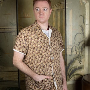 Peut inclure: Une chemise à manches courtes avec un motif floral marron et beige. La chemise est portée sur un t-shirt blanc et associée à un pantalon gris. La chemise a une coupe décontractée et une poche sur le côté gauche.