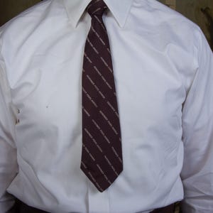 May include: A white dress shirt with a dark burgundy tie. The tie has a diagonal pattern of text in a lighter color. The shirt is buttoned at the collar and the sleeves are long.