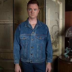 May include: A medium-wash denim jacket with button closures and two chest pockets. The jacket is worn with matching denim pants. The person is standing in front of a vintage-style screen.