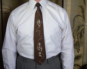 May include: A brown tie with a repeating pattern of stylized figures in white and blue, worn with a white dress shirt and gray trousers. The tie features a unique design, adding a touch of vintage flair to the outfit.