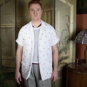 Peut inclure: Une chemise blanche à manches courtes avec de petits motifs floraux rouges, portée sur un t-shirt gris et associée à un pantalon gris. La chemise a deux poches avant et une coupe décontractée. La personne se tient devant un paravent décoratif.