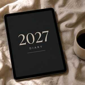 May include: A black tablet screen displaying the text 2027 DIARY in a light beige serif font, accompanied by a small white ceramic mug filled with dark liquid, positioned on a soft, off-white fabric surface with subtle shadows.