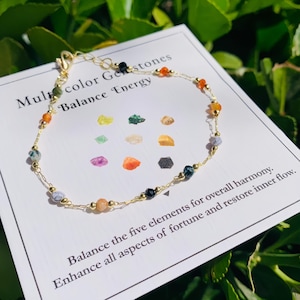 May include: A delicate gold chain bracelet adorned with multicolored gemstones. The bracelet features a variety of stone colors, including orange, green, black, and white. The bracelet is displayed on a white card with text about balance and energy.