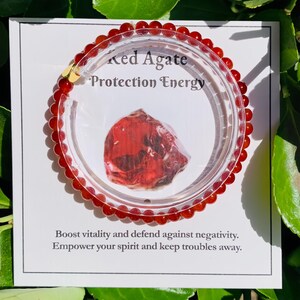 May include: A red agate bead bracelet with a clear plastic display. The display card reads "Red Agate Protection Energy." The bracelet is made of small, round, red beads. The card also includes text about boosting vitality and defending against negativity.