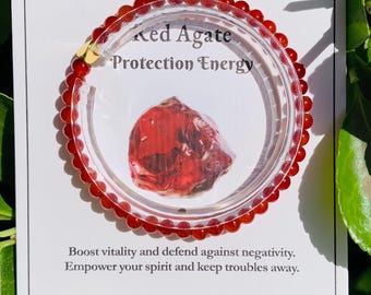 May include: A red agate bead bracelet with a clear plastic display. The display card reads "Red Agate Protection Energy." The bracelet is made of small, round, red beads. The card also includes text about boosting vitality and defending against negativity.