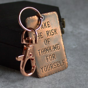 Puede incluir: Un llavero de color cobre con un anillo y una etiqueta que dice "Toma el riesgo de pensar por ti mismo".