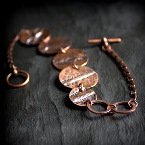 May include: A copper bracelet featuring a chain and five round, hammered copper discs. The discs are linked together and to the chain with small copper rings.