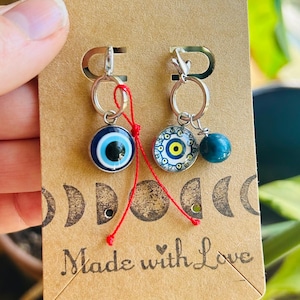 May include: A pair of silver earrings with blue and white evil eye charms and a blue bead. The earrings are attached to a brown card with the words "Made with Love" printed on it. The card has a crescent moon design.