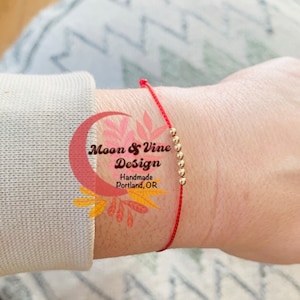 May include: Red string bracelet with gold beads. The bracelet is tied around a person's wrist. The text "Moon & Vine Design Handmade Portland, OR" is visible in the image.