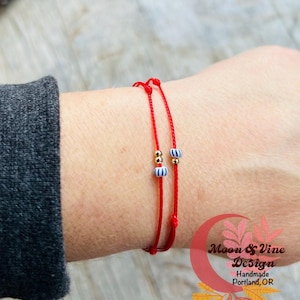 May include: Two red string bracelets with gold and blue striped beads. The bracelets are tied together and worn on a wrist. The text "Moon & Vine Design Handmade Portland, OR" is visible in the lower right corner of the image.