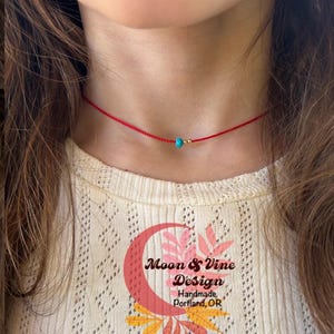 May include: A delicate red choker necklace featuring a small turquoise bead and gold accents. The necklace is worn around the neck. The image also shows a cream-colored top and the logo for Moon & Vine Design.