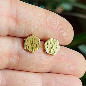 May include: A pair of gold-colored hexagon stud earrings. The earrings have a textured surface and a simple, modern design. The earrings are small and delicate, suitable for everyday wear.