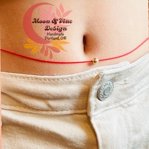 May include: A red string tied around a person's waist with a small gold bead in the center. The string is tied in a knot at the front and the bead is centered on the knot. The image also includes a logo for Moon & Vine Design, Handmade Portland, OR.