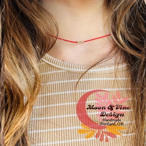 May include: A red string choker necklace with small gold beads. The necklace is worn by a person with long brown hair. The background is a tan and white striped shirt with the text "Moon & Vine Design Handmade Portland, OR".