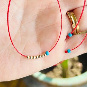 May include: A delicate red string bracelet with small gold beads and turquoise accents. The bracelet is shown against a blurred background, highlighting the jewelry's fine details and craftsmanship. The bracelet is a simple yet elegant accessory.