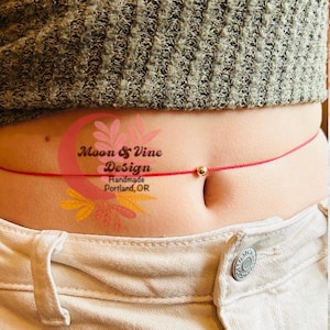 May include: A red string tied around a person's waist with a small gold bead in the center. The string is tied in a knot at the front and the bead is centered on the knot. The text "Moon & Vine Design Handmade Portland, OR" is visible on the person's stomach.