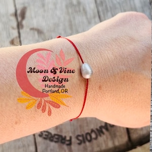 May include: A red string bracelet with a silver bead charm. The bracelet is on a person's wrist. The text "Moon & Vine Design Handmade Portland, OR" is visible on the person's arm.