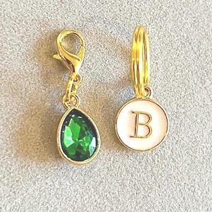May include: Two gold-tone charms, one with a green teardrop-shaped crystal and the other with a white enamel circle with the letter "B" in gold.