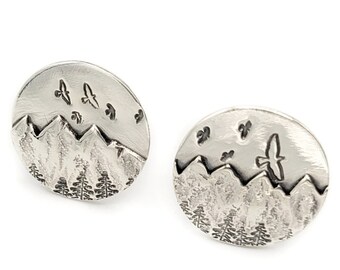 May include: Two silver-toned stud earrings. Each earring features a mountain range, trees, and flying birds. The earrings have a circular shape and a detailed, etched design. The overall theme is nature and outdoors.