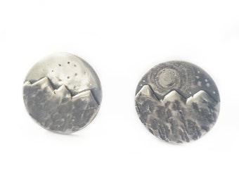 May include: A pair of silver earrings with a mountain range design. The earrings are round and have a textured surface. One earring features a starry sky above the mountains, while the other earring features a moon and stars.