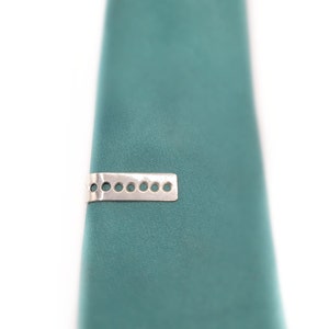 May include: Silver tie clip with a rectangular design and multiple circular cutouts. The tie clip is on a teal tie.