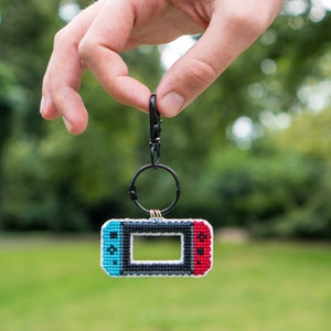 May include: A black keychain with a blue, red, and gray cross-stitch design of a Nintendo Switch console.