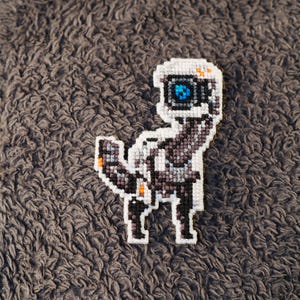 Puede incluir: Un robot pixelado blanco y gris con un ojo azul, bordado en una tela marrón y peluda.