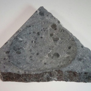 May include: A gray stone soap dish with a rounded, slightly indented center. The dish has a rough, textured surface with dark gray speckles.