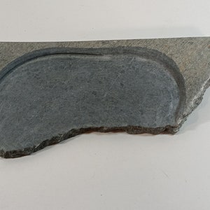 May include: A gray slate stone with a carved out oval shape in the center. The stone has a rough, uneven edge.